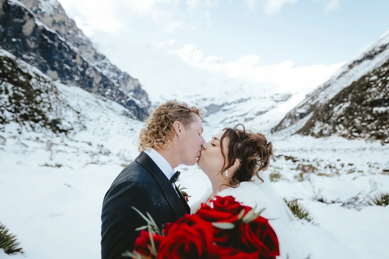 Couple kissing with a bouquet of red roses in the snow-filled Earnslaw Burn valley during a winter heli elopement