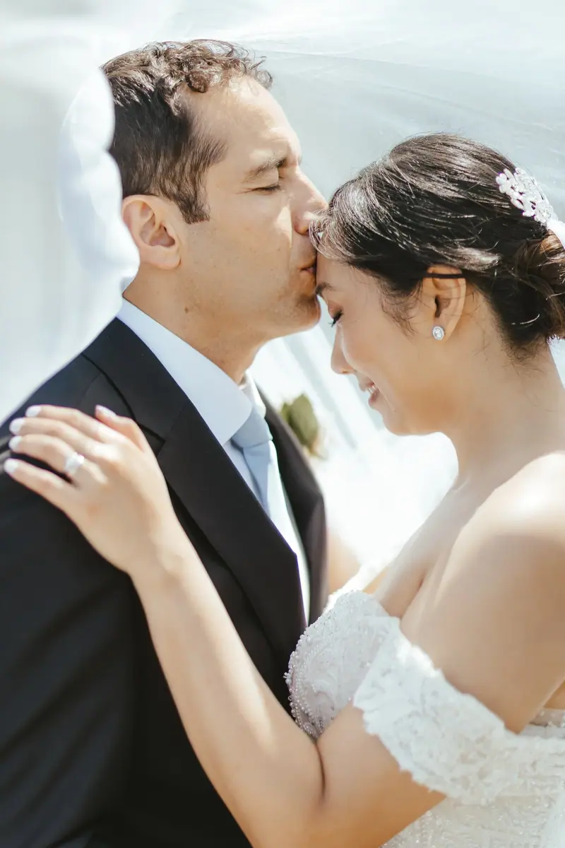 A bride and groom sharing a kiss during their wedding, with the groom in a black tuxedo and the bride in a white lace wedding dress, her hair adorned with a decorative hairpiece.