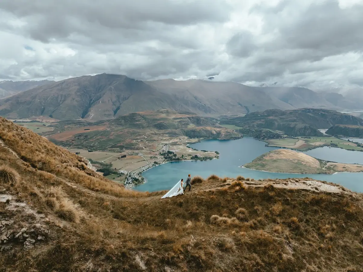 A bride and groom holding hands on a hillside with a lake and mountains in the background, under cloudy skies.