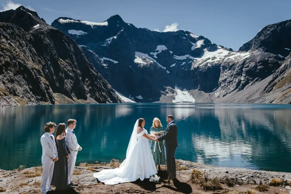 A wedding ceremony taking place outdoors by a lake, with mountains and snow patches in the background. The bride and groom are holding hands, and a officiant is reading from a book. Two of the wedding party members stand to the left.