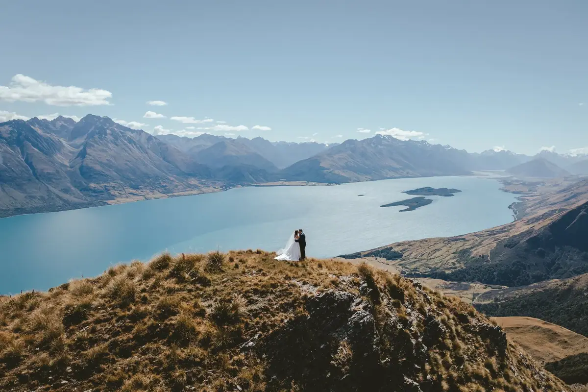 A bride and groom sharing a kiss on a hilltop overlooking a large lake, mountains, and a clear blue sky.
