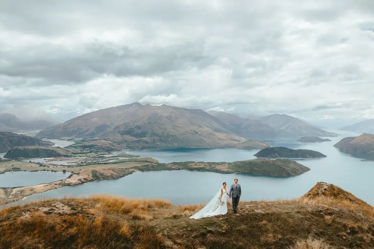 A newlywed couple standing on a hill with a scenic lake and mountain landscape in the background, under a cloudy sky.