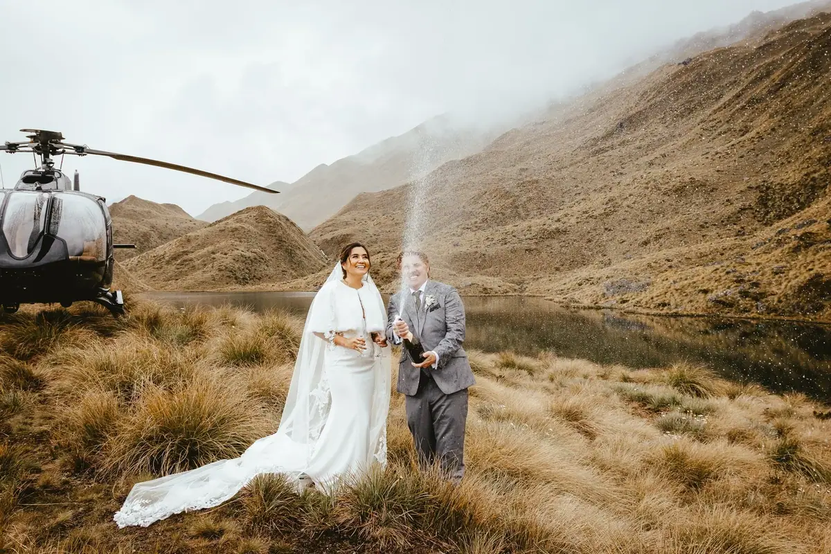 A bride and groom celebrating outdoors near a lake with mountains in the background, a helicopter with a camera is nearby, and the groom is opening a bottle of champagne.