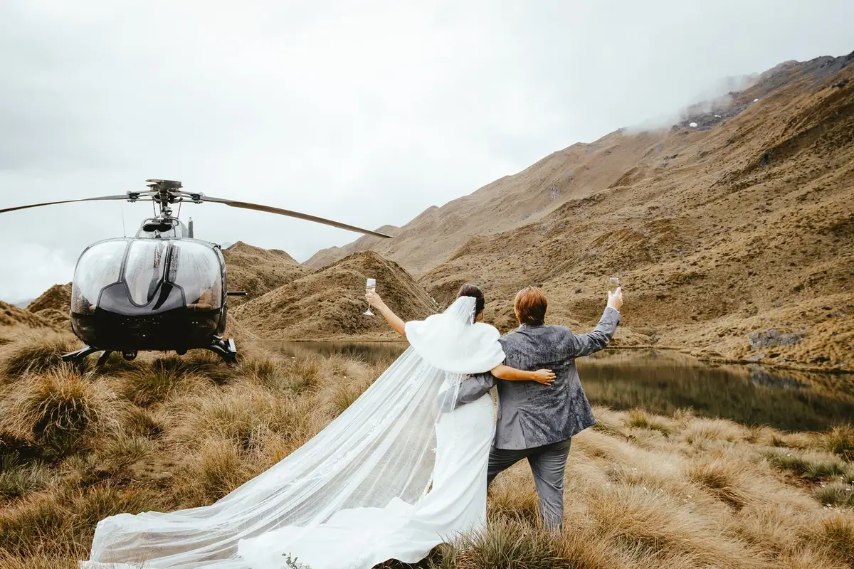 A bride and groom in wedding attire sitting on the grass beside a lake, holding glasses of champagne, with a helicopter parked nearby in a mountainous landscape.