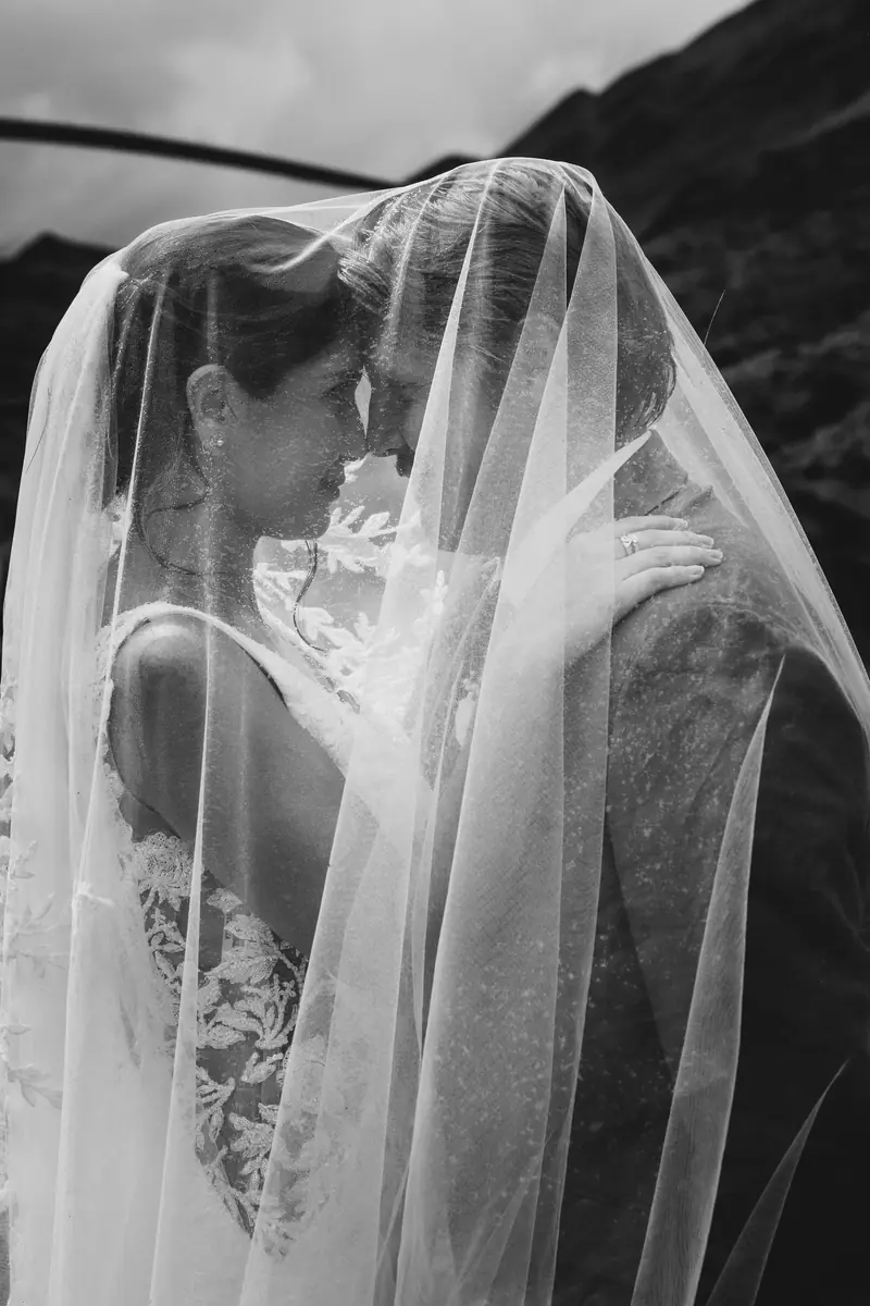 A bride with short hair wearing a lace wedding dress and veil embraces a person outdoors with mountainous terrain in the background.