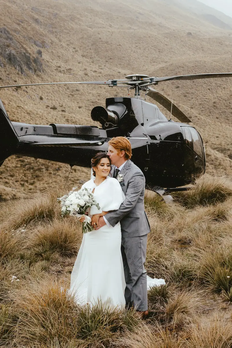 A newlywed couple standing in a grassy field with a helicopter in the background on a mountain hillside.
