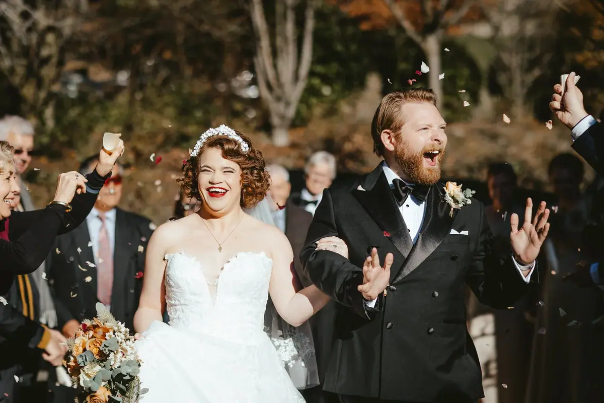 A bride and groom celebrating their wedding outdoors with guests throwing flower petals. The bride wears a white wedding dress and veil, holding a bouquet, and has curly hair with a floral headpiece. The groom is in a black tuxedo with a bow tie and