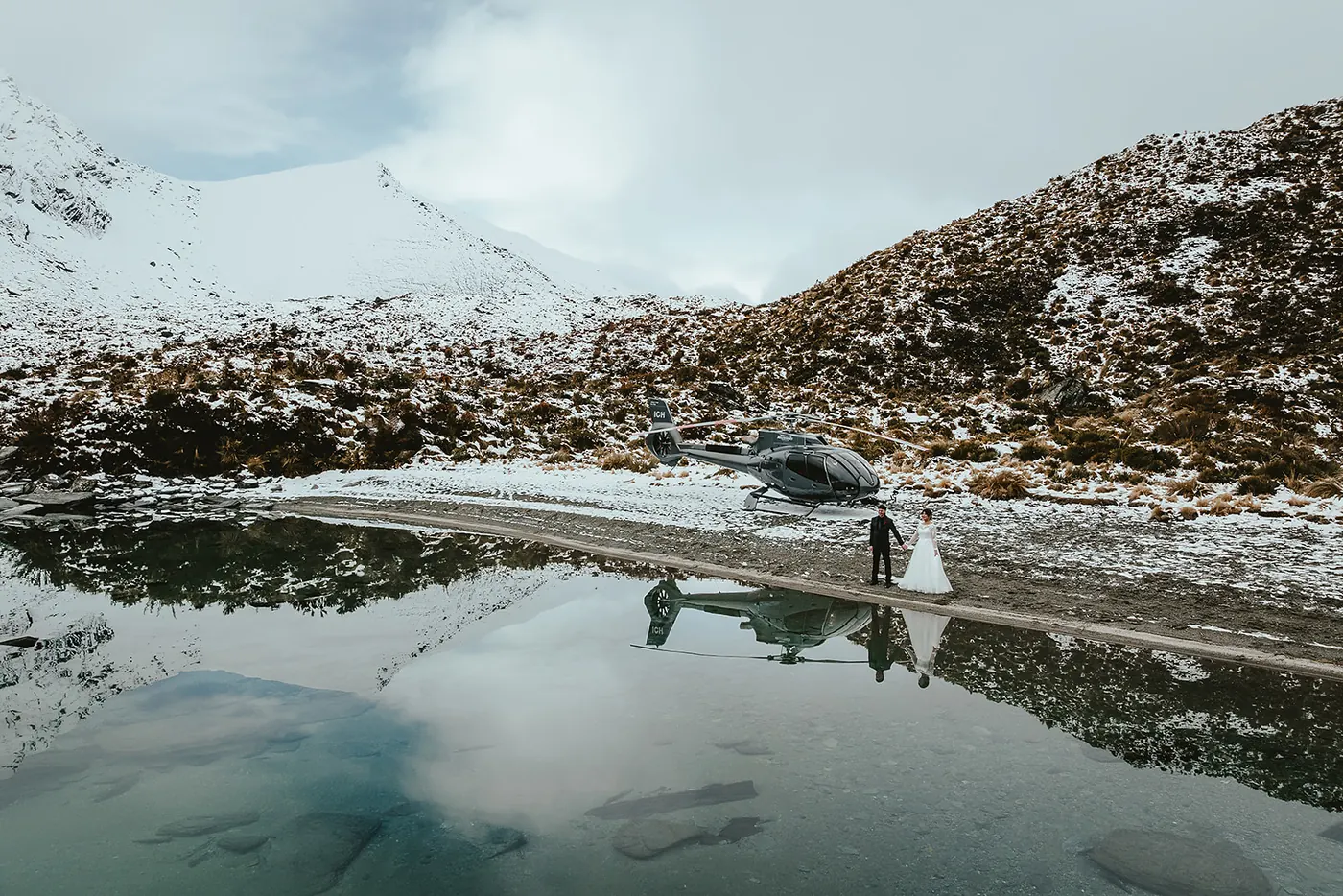 Couple holding hands beside their helicopter, reflected in a still snowy alpine lake on a heli elopement.