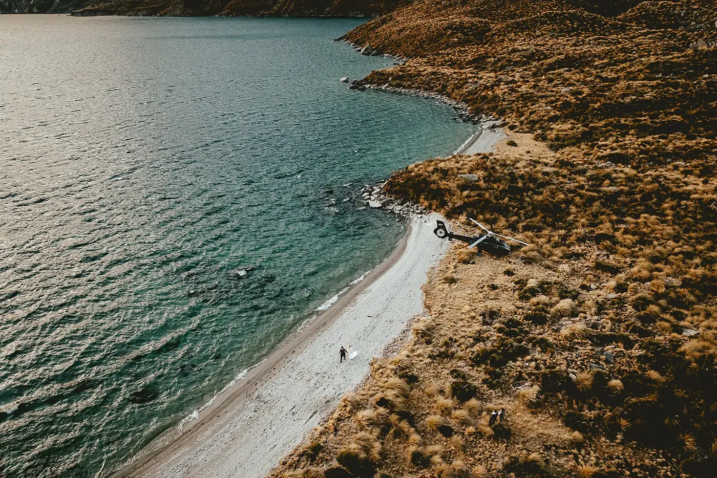 Aerial view of a bride and groom walking a pale Lake Lochnagar beach, their helicopter landed in the golden tussock beside turquoise water