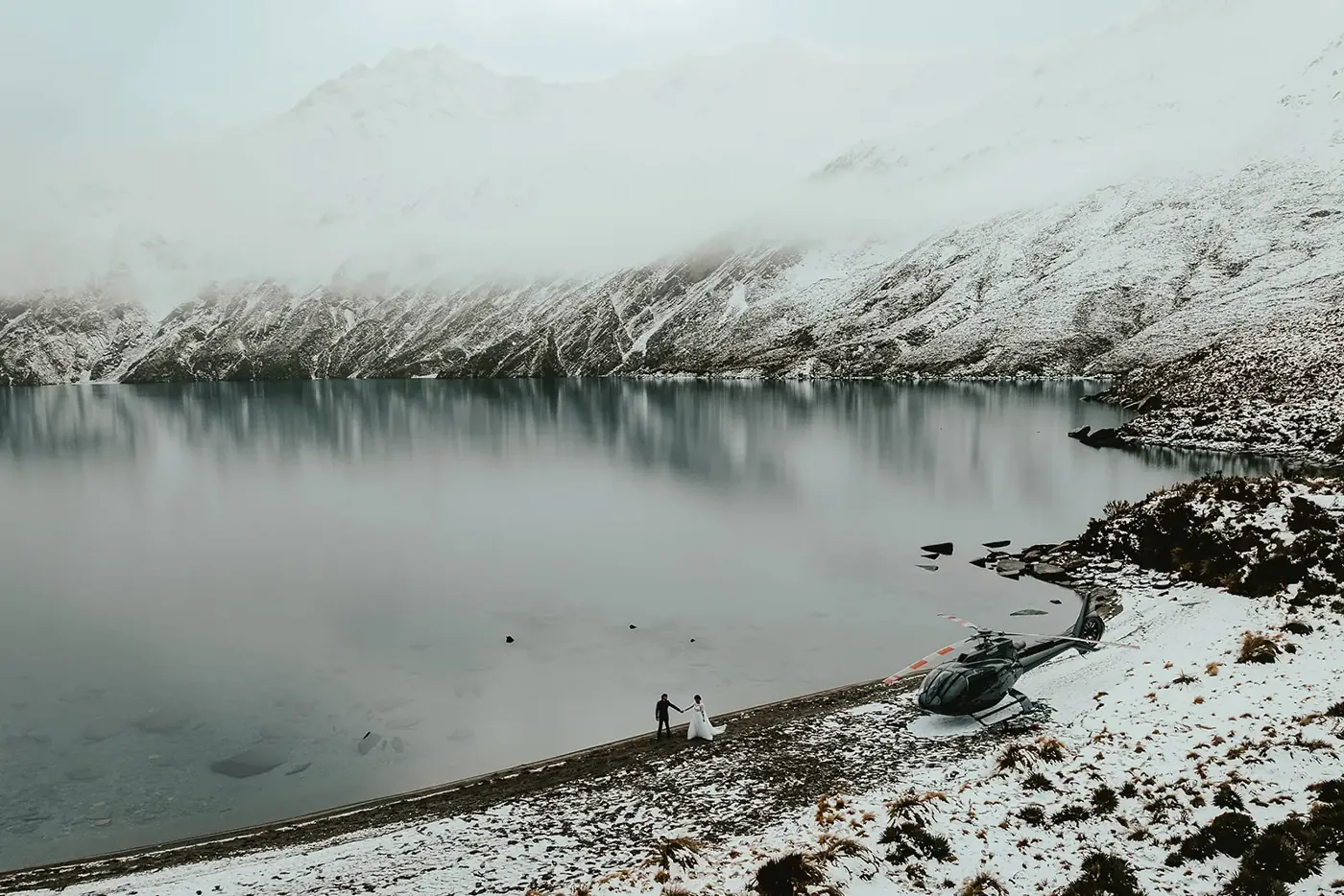 Aerial view of a helicopter and couple on the snowy shoreline of Lake Lochnagar beneath cloud-wrapped peaks