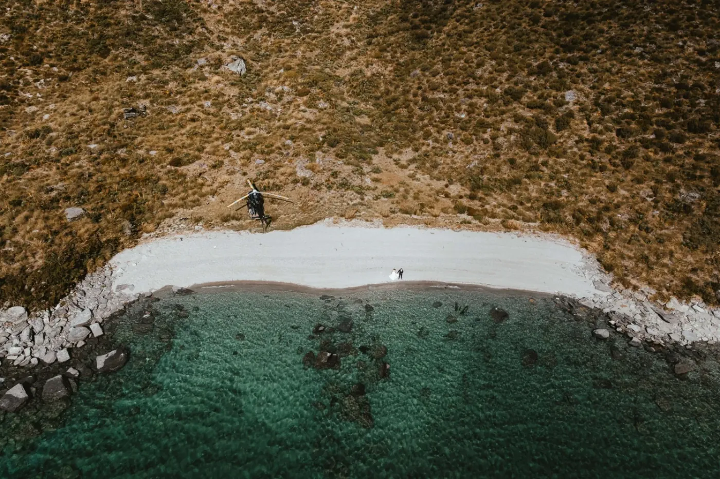 Aerial view of a helicopter landed above a pale Lake Lochnagar beach with a bride and groom by the clear green water