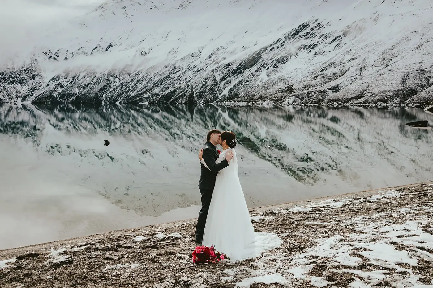 Bride and groom embracing on the shore of Lake Lochnagar as snowy mountains reflect in the still water