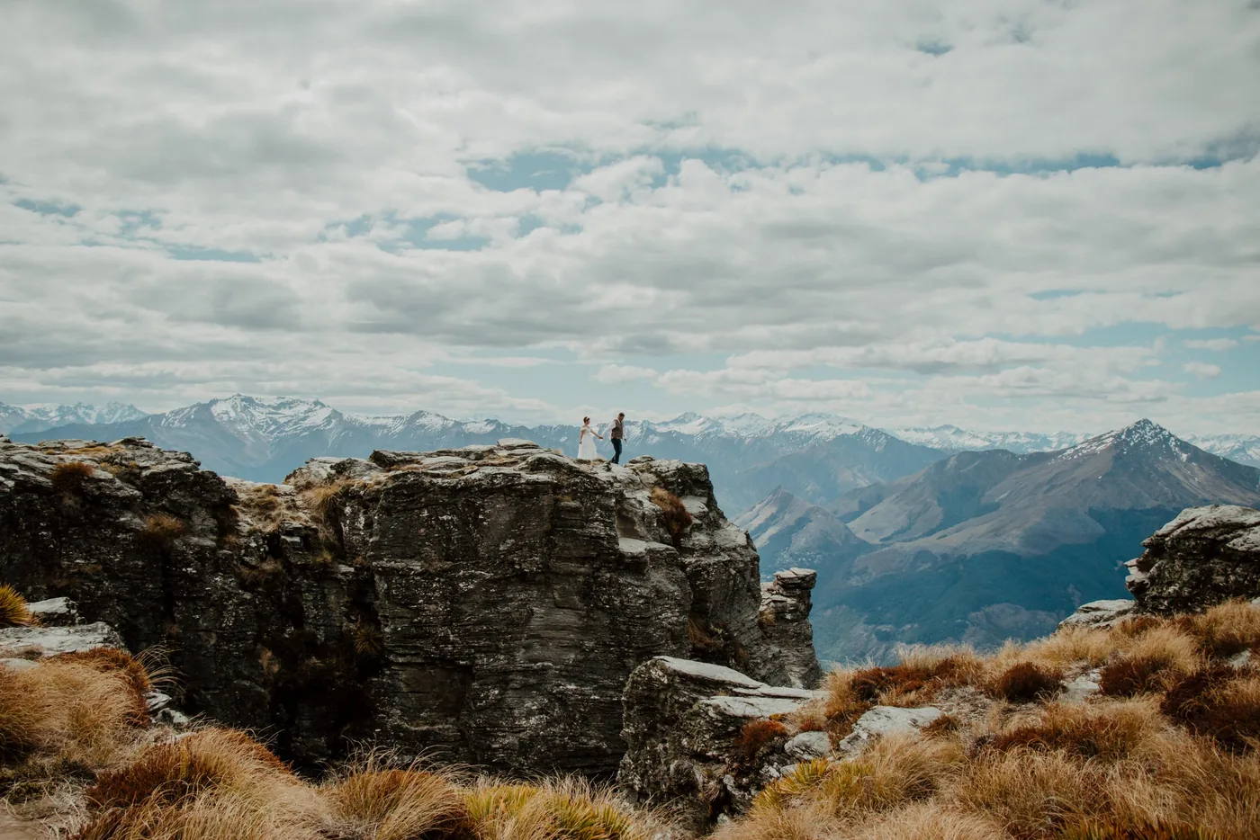 A couple standing together on a rocky alpine ridge above Queenstown with snow-capped peaks behind them.