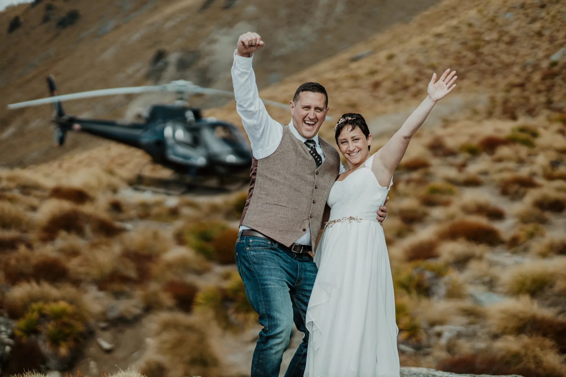 Couple celebrating at Lower Cecil Peak with their helicopter on the tussock behind them, Queenstown.