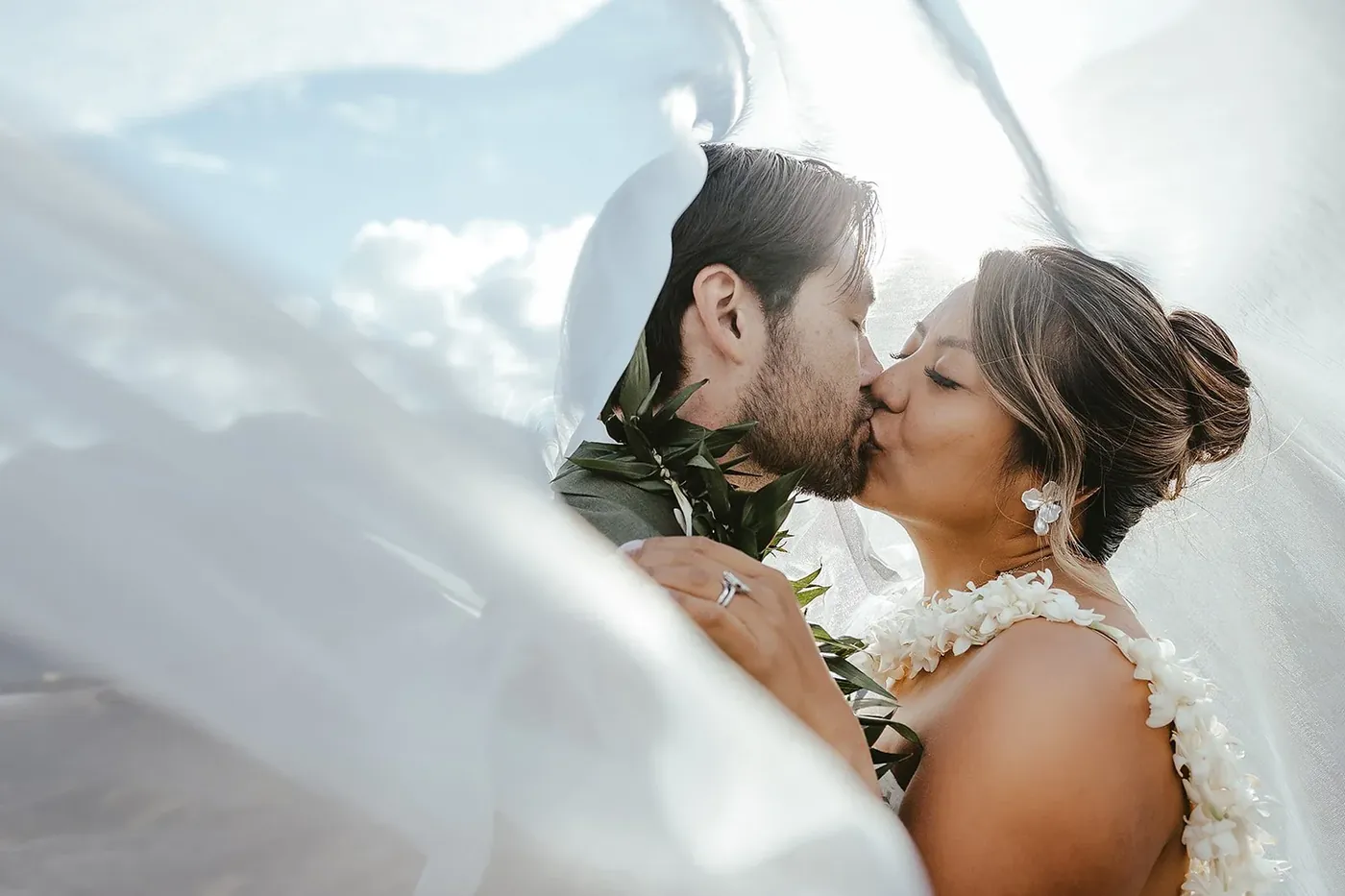 A wedding couple sharing a kiss under a veil with blue sky and clouds.